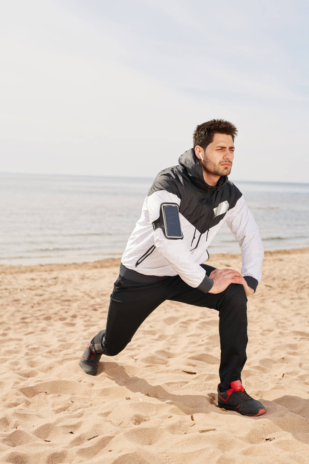 Young sportsman with airpods and smartphone exercising on sandy beach with waterside on background